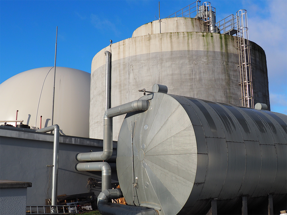 Große zylindrische und kugelförmige Behälter einer Biogasanlage mit sichtbaren Rohren und Leitern unter blauem Himmel.