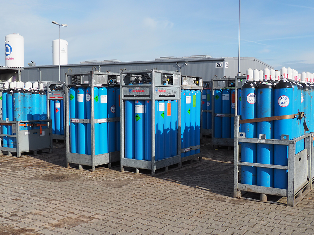 Mehrere blaue Gasflaschen in Metallrahmen auf einem gepflasterten Hof vor Industriegebäuden unter blauem Himmel.