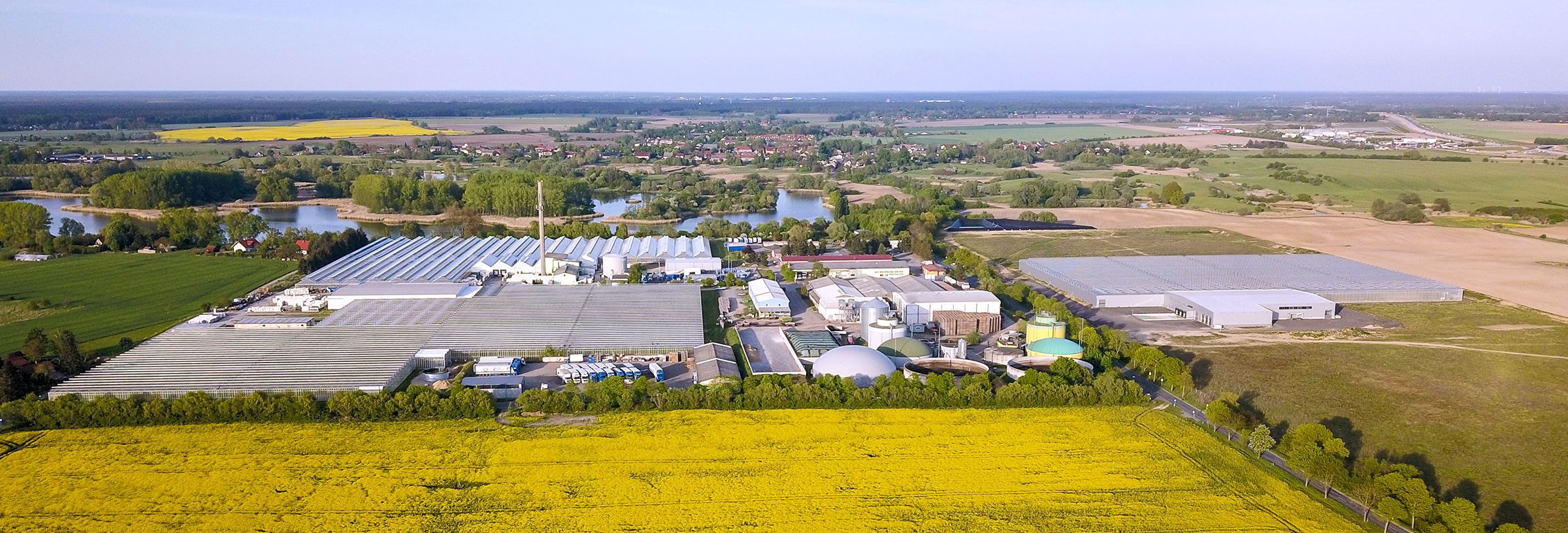 Luftaufnahme einer Industrieanlage mit Gewächshäusern, Biogasanlage und gelbem Rapsfeld im Vordergrund, Dorf und Felder im Hintergrund.