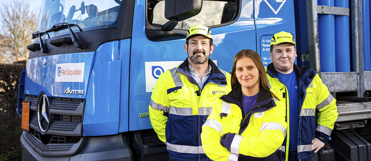 header_bilder_webseite_Karriere_ausbildung_kategorie_1200x521px Drei Personen in gelb-blauen Warnjacken stehen vor einem blauen Mercedes-Benz Lkw mit Aufschrift 'Air Liquide'.
