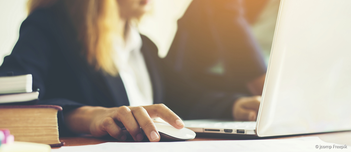 header_bilder_webseite_Karriere_verwaltung_intro_1200x521px Frau in Businesskleidung arbeitet an einem Laptop, Hand auf weißer Computermaus, Bücher und Papiere auf dem Tisch.