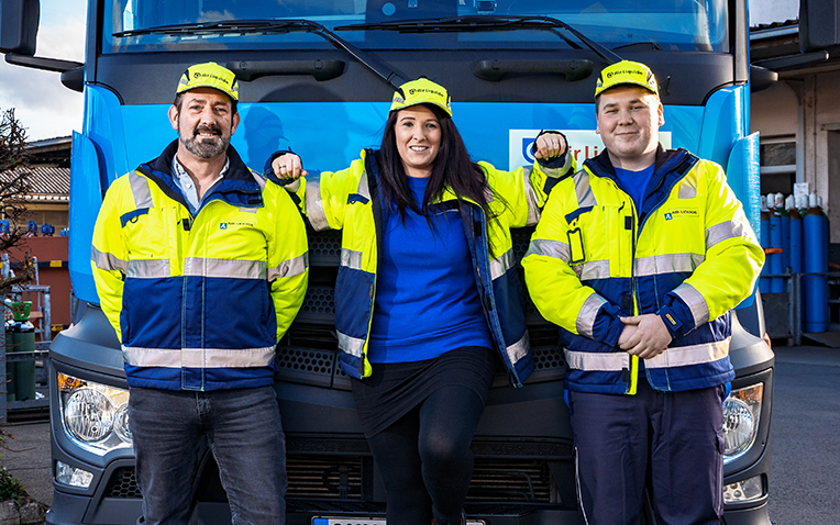 Drei Personen in gelb-blauen Warnjacken und gelben Mützen lehnen vor einem blauen Lkw mit deutschem Kennzeichen.