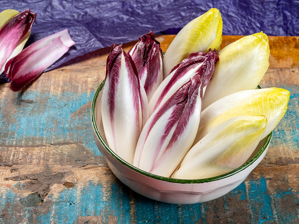 Schale mit mehreren roten und weißen Chicorée-Salaten auf rustikalem Holzuntergrund, zwei Chicorée liegen daneben.