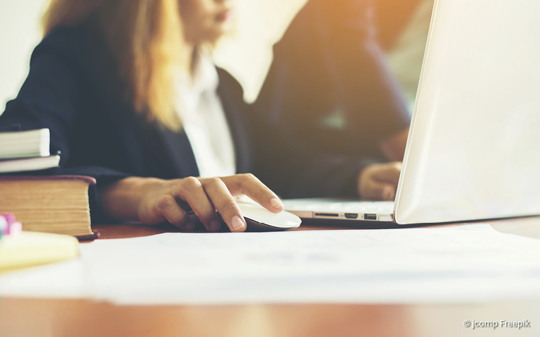 Frau in Businesskleidung arbeitet an einem Laptop, Hand auf weißer Computermaus, Bücher und Papiere auf dem Tisch.