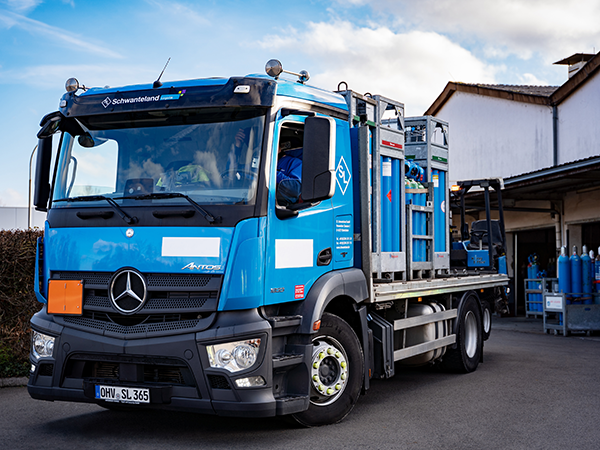 Blauer Mercedes-Benz Lkw mit "Schwanteland"-Schriftzug auf der Fahrerkabine, beladen mit Gasflaschen, steht vor einem Gebäude.