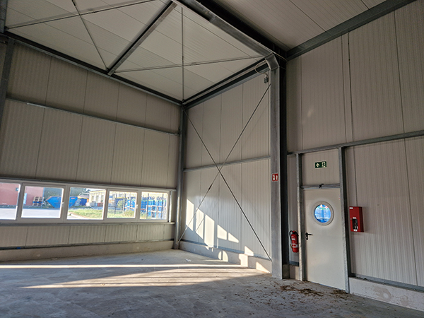 Leerer Lagerraum mit großen Fenstern, einer weißen Tür mit rundem Fenster, Feuerlöscher und Notausgangsschild.