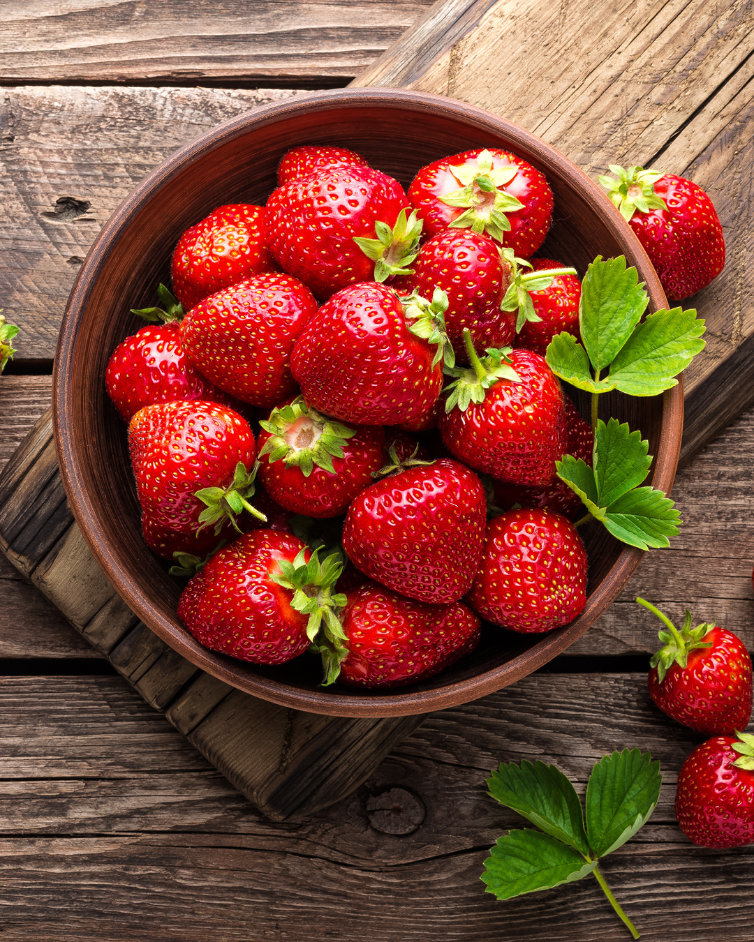 Braune Schale mit mehreren frischen, roten Erdbeeren und grünen Blättern auf Holzuntergrund.
