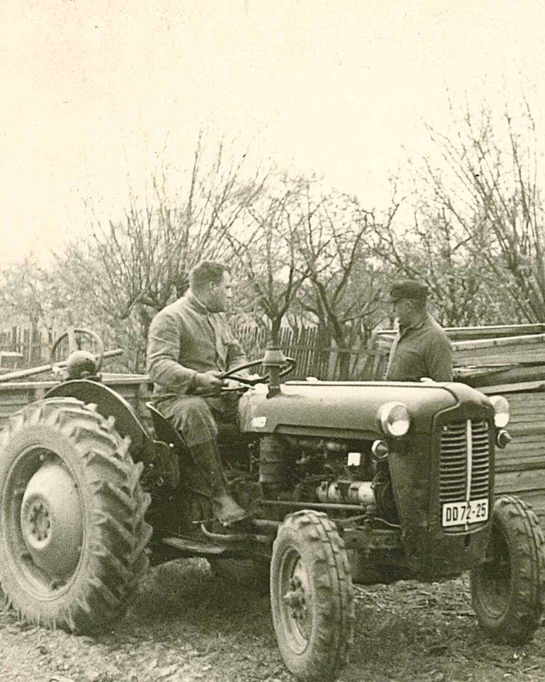 Alter Traktor mit zwei sitzenden Personen, im Hintergrund Bäume und Holzzaun.
