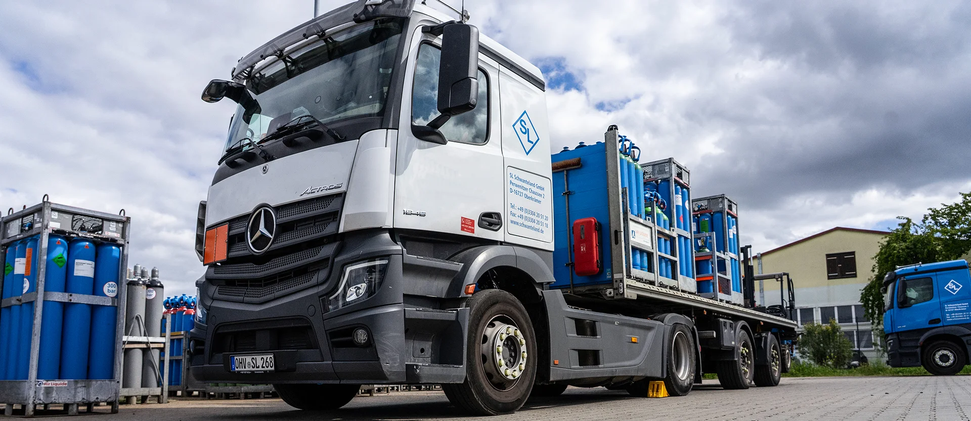 Lastwagen mit Anhänger, beladen mit Gasflaschen, steht auf einem gepflasterten Hof neben weiteren Gasflaschen und einem weiteren Lastwagen im Hintergrund